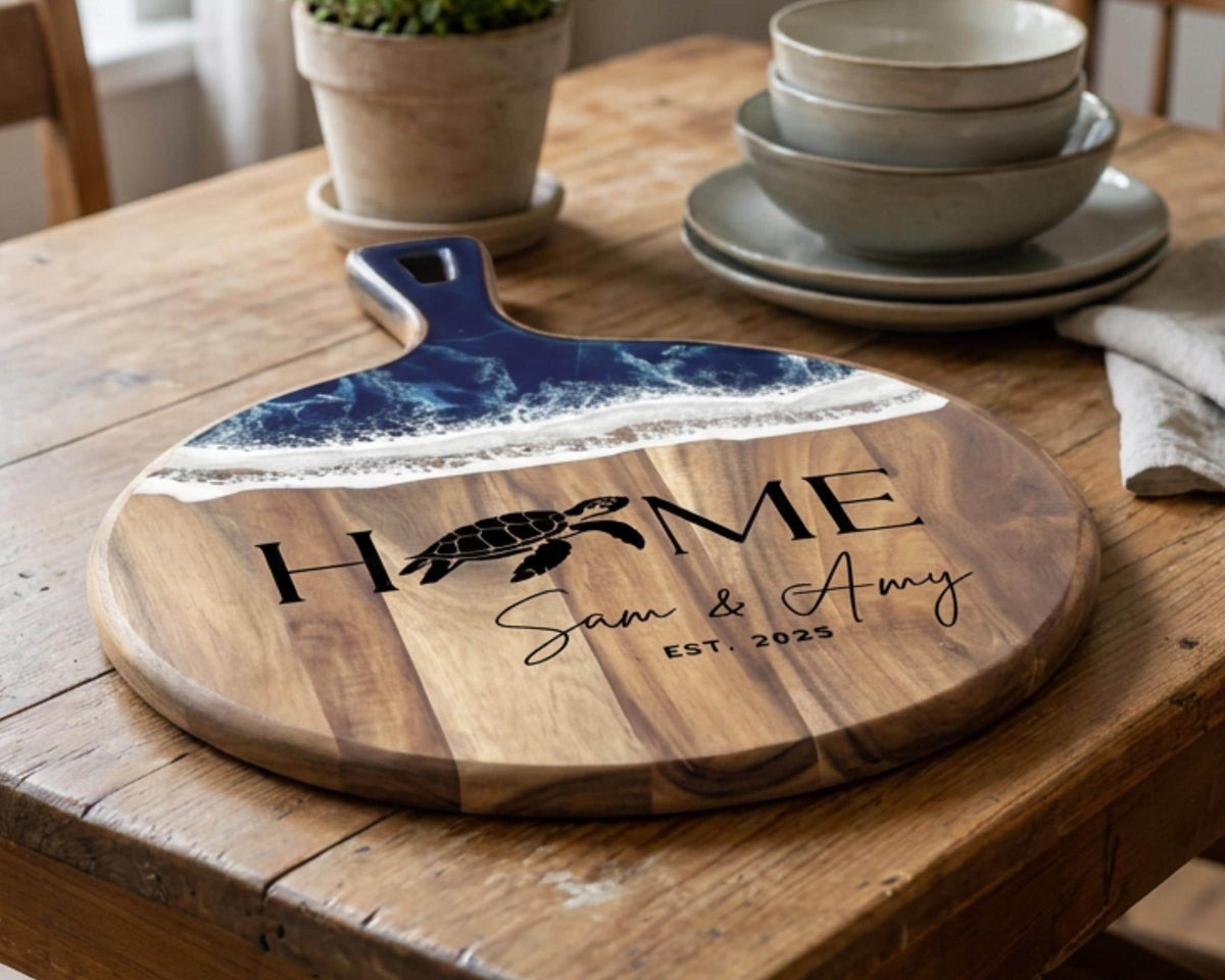 A wooden cutting board with a blue and white wave design and the text "HOME Sam & Amy EST. 2025" printed on it, along with a turtle image. The cutting board is placed on a wooden table, and there are bowls and a potted plant nearby.
