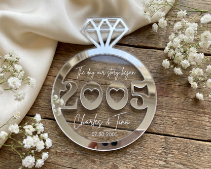A clear glass ornament with the numbers "2005" and the names "Charles & Tina" written on it, placed on a wooden surface surrounded by white flowers.