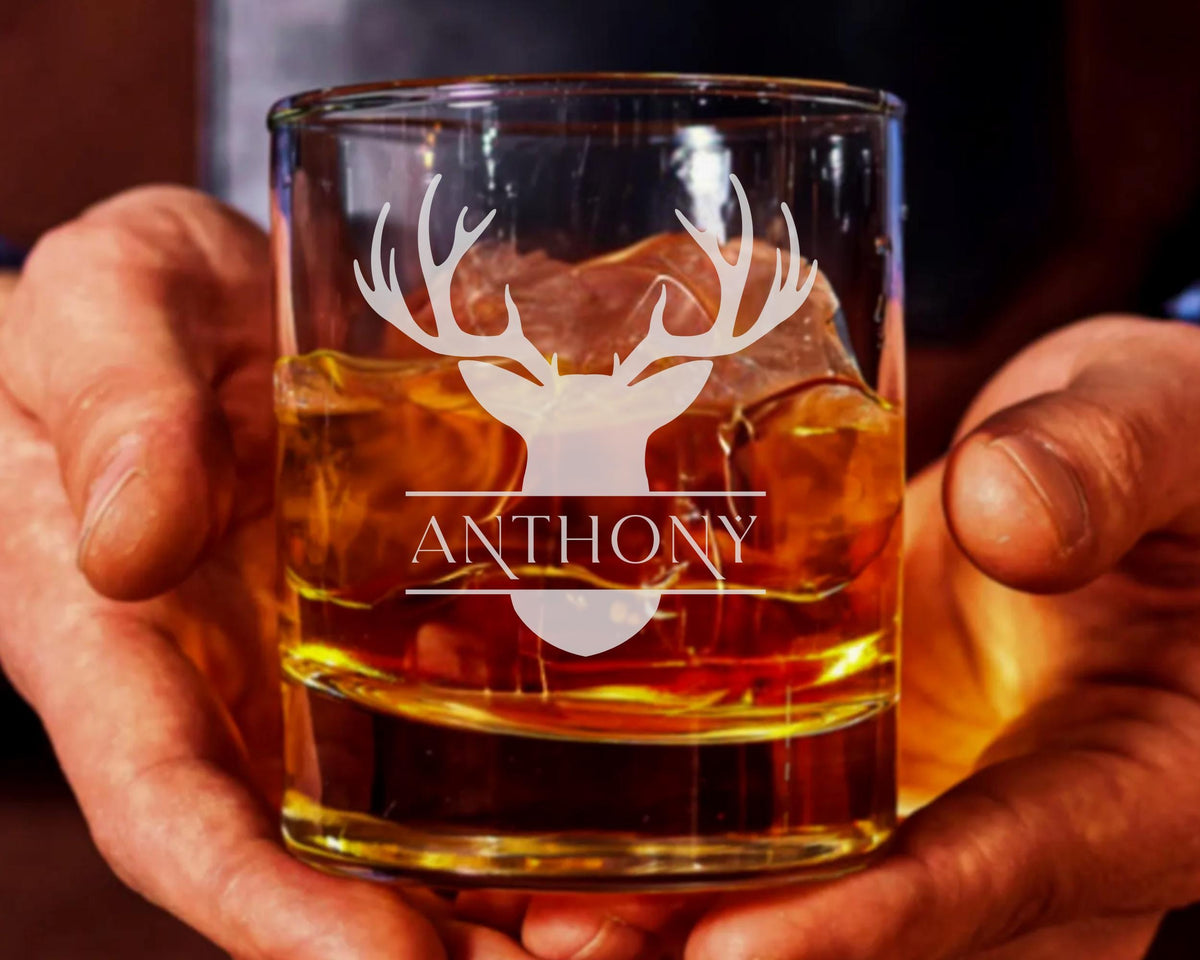 A person's hands holding a glass filled with amber-colored liquid, likely whiskey, with a deer head silhouette printed on it.