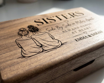 A wooden box with a carved design featuring two women sitting together, accompanied by the text "Sisters Side By Side Or Miles Apart Always Close At Heart Emma & Sarah".