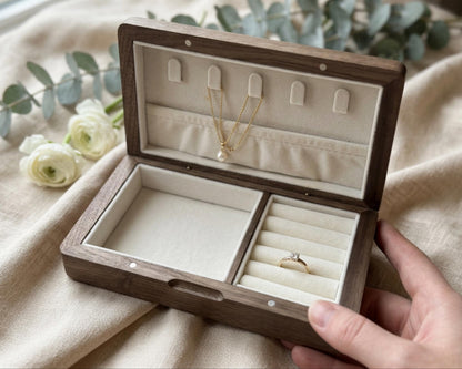 A wooden jewelry box with a compartment for storing a ring and a compartment for displaying a necklace. The box is placed on a beige cloth, and there are white roses in the background.