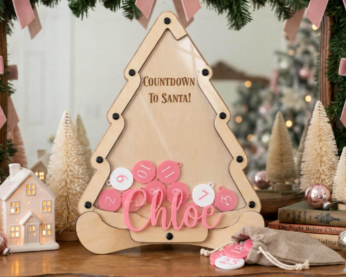 A wooden countdown clock with the text "Countdown To Santa!" and the name " Chloe" written on it, surrounded by pink and white ornaments and a small Christmas tree.