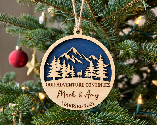 A wooden ornament hanging on a Christmas tree, featuring a mountain landscape and the text "OUR ADVENTURE CONTINUES Mark & Amy MARRIED 2001".
