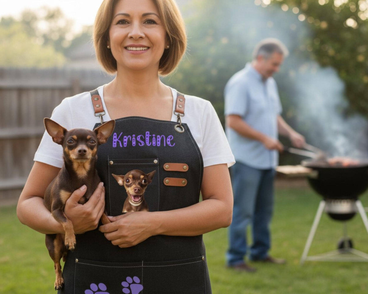 A woman wearing a black apron with a dog's face and name on it is holding a small dog in her arms. In the background, a man is grilling food on a barbecue.