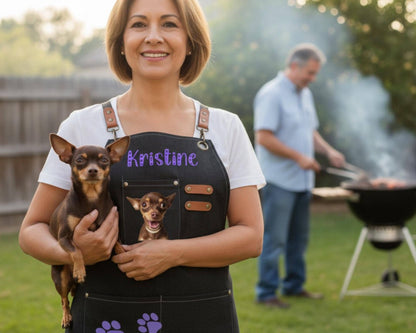 A woman wearing a black apron with a dog's face and name on it is holding a small dog in her arms. In the background, a man is grilling food on a barbecue.