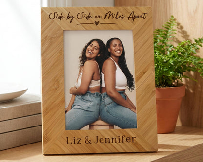 A wooden picture frame with two smiling women posing together, placed on a wooden surface next to a potted plant.