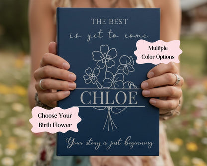 A person is holding a blue book with the title "The Best is yet to Come" and the name " Chloe" printed on it. The book also has a section titled "Choose Your Birth Flower" and a section titled "Your story is just beginning".