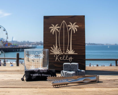 a wooden sign with a surfboard design, a glass, and some grey stones on a wooden table, with a ferris wheel and a body of water in the background.