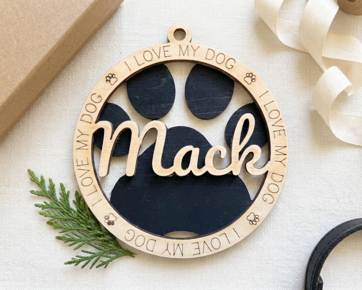 A wooden ornament with the name "Mac" engraved on it, surrounded by a circular frame with paw prints and the phrase "I LOVE MY DOG" written around the edges.