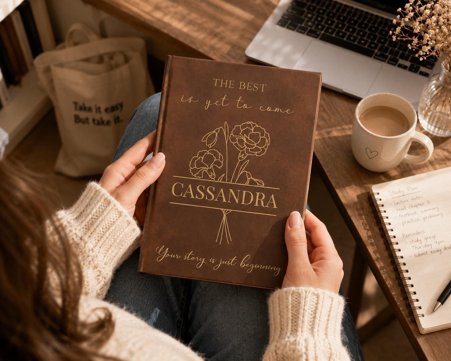 A person is sitting at a desk, holding a book titled "CASSANDRA" with a floral design on the cover. The desk also has a laptop, a coffee cup, and a notebook.