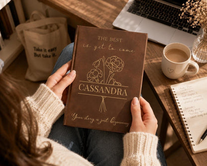 A person is sitting at a desk, holding a book titled "CASSANDRA" with a floral design on the cover. The desk also has a laptop, a coffee cup, and a notebook.