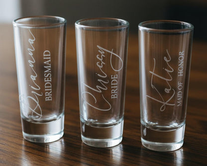 Three shot glasses with personalized names printed on them, placed on a wooden surface.