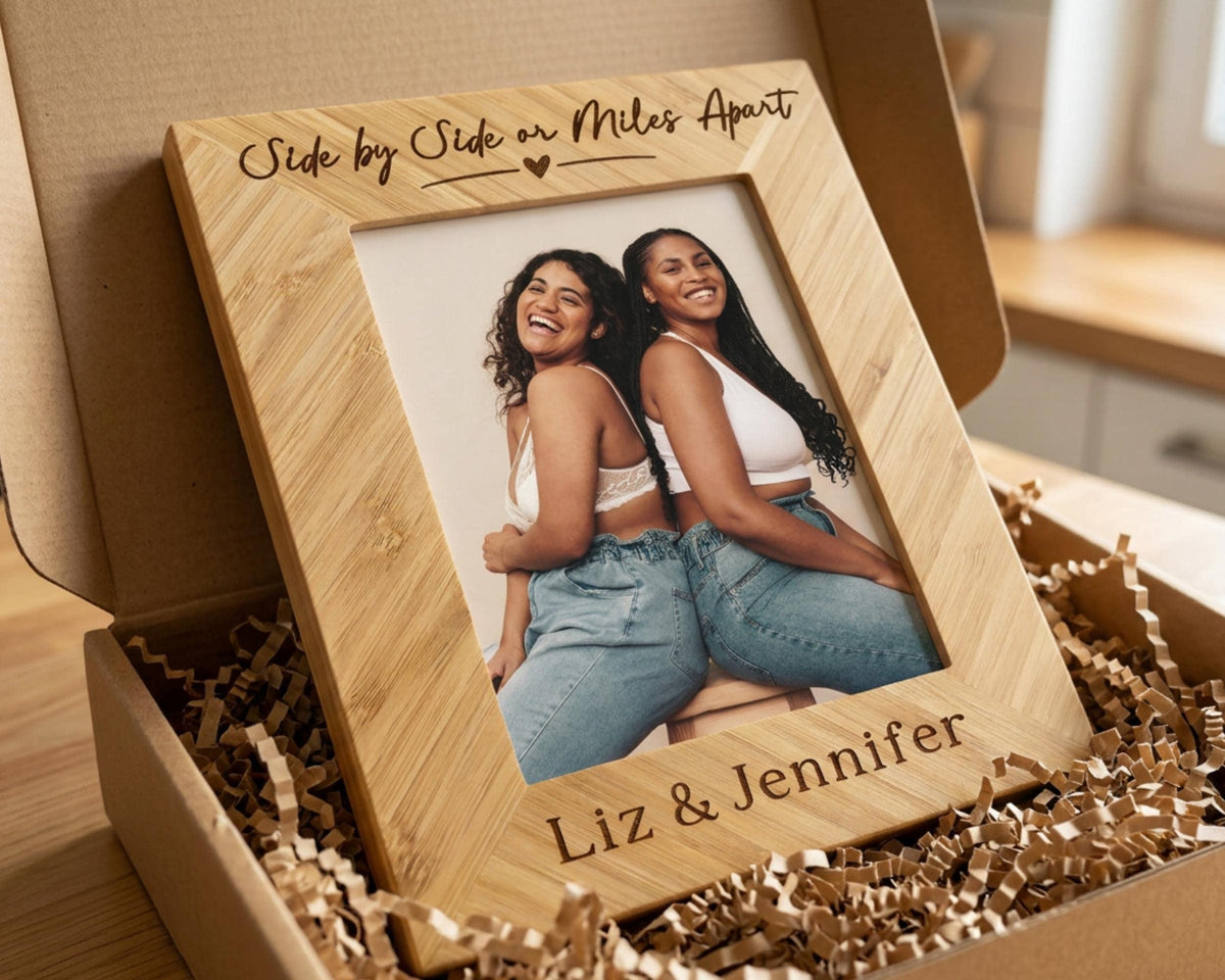 A wooden picture frame with a photo of two smiling women inside, placed in a cardboard box filled with shredded paper.