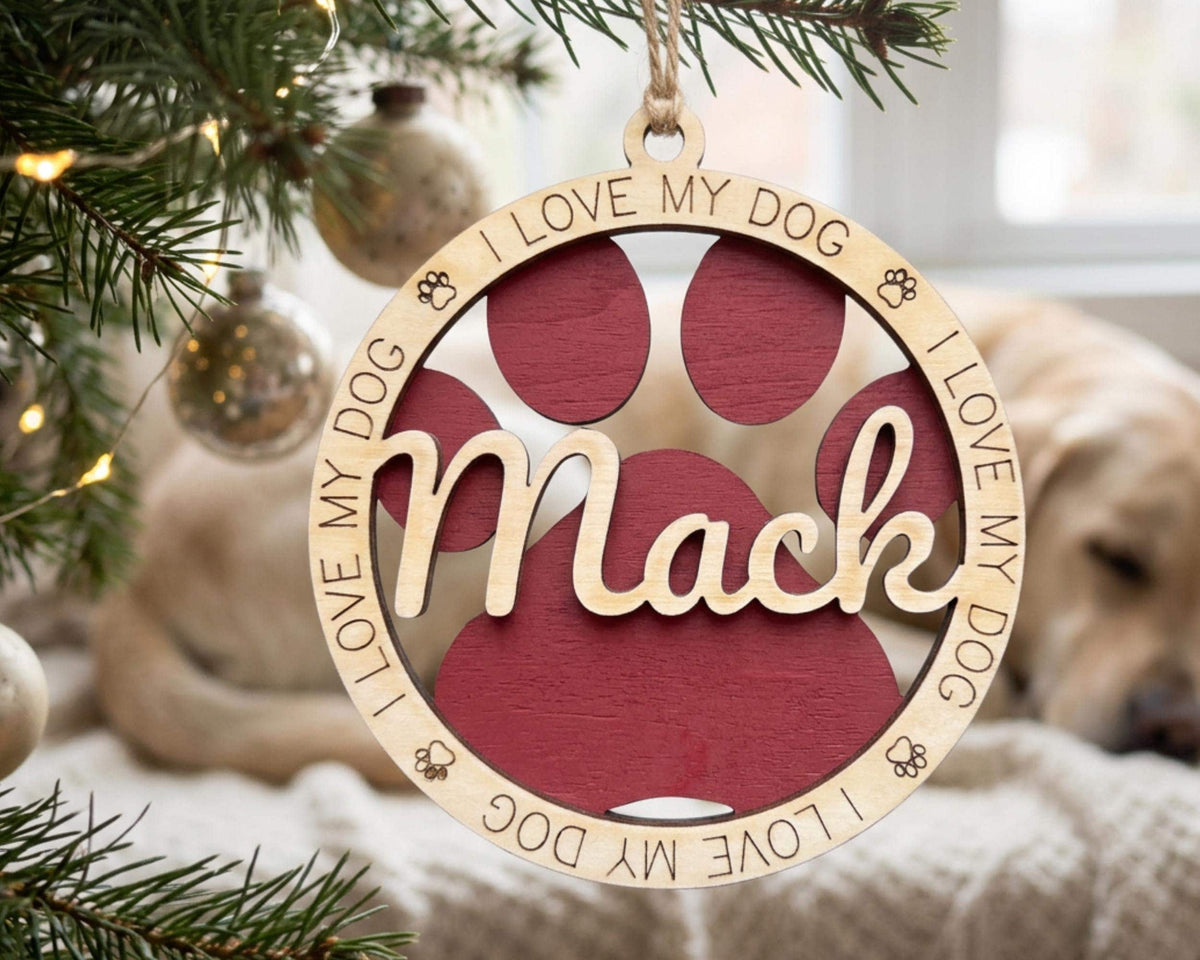 A wooden ornament with a red paw print and the name "Mac" engraved on it, hanging on a Christmas tree.