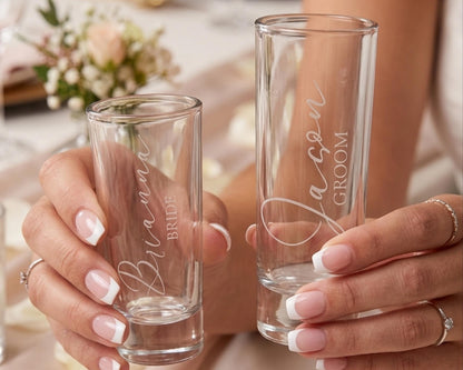 A person's hands holding two personalized shot glasses with names engraved on them.