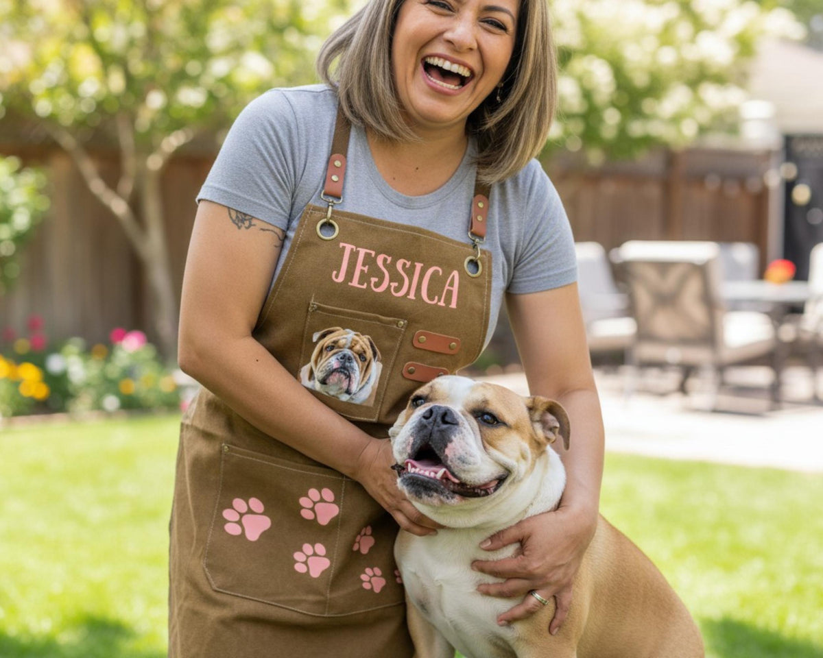 A woman wearing a brown apron with the name "JESSICA" and a picture of a bulldog is smiling and holding a brown and white bulldog in a grassy outdoor setting.