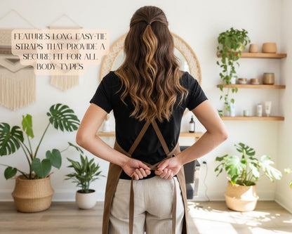 A woman with long, wavy brown hair stands in a room with potted plants, wearing a black shirt and a brown apron. The text overlay on the image reads "FEATURES LONG EASY-TIE Straps that provide a secure fit for all body types".