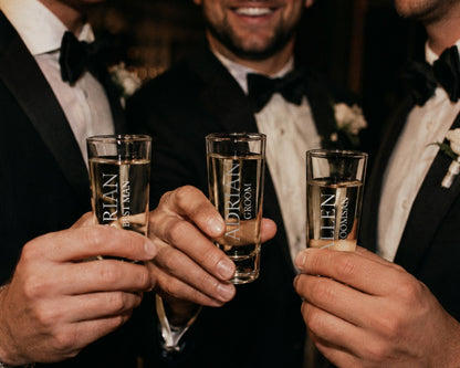 Three men in tuxedos are holding up shot glasses, each with a personalized name printed on it, in a celebratory toast.