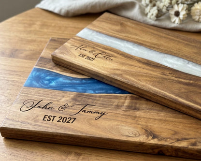 Two personalized acacia cutting boards with blue pearl resin rivers and engraved couple names