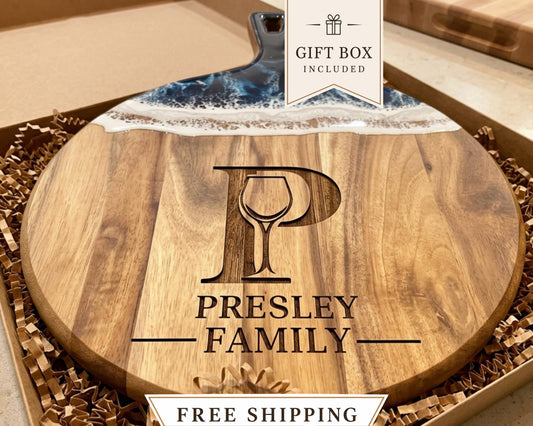 A wooden cutting board with a personalized monogrammed logo, including a wine glass and the text "PRESLEY FAMILY". The cutting board is presented in a gift box, and there is a "GIFT BOX INCLUDED" label on the top right corner.