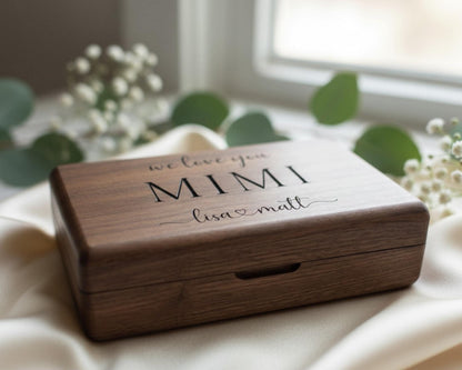 A wooden box with the text "we love you Mimi Lisa & Matt" engraved on it, placed on a white cloth near some greenery.