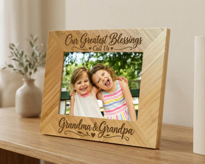 A wooden picture frame with a photo of two young girls inside, placed on a wooden surface. The frame has a decorative border and the text "Our Greatest Blessings Call Us" written on it.