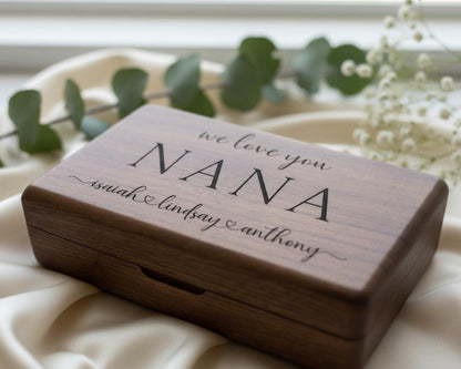 A wooden box with the inscription "we love you NANA" engraved on it, placed on a white cloth with greenery in the background.