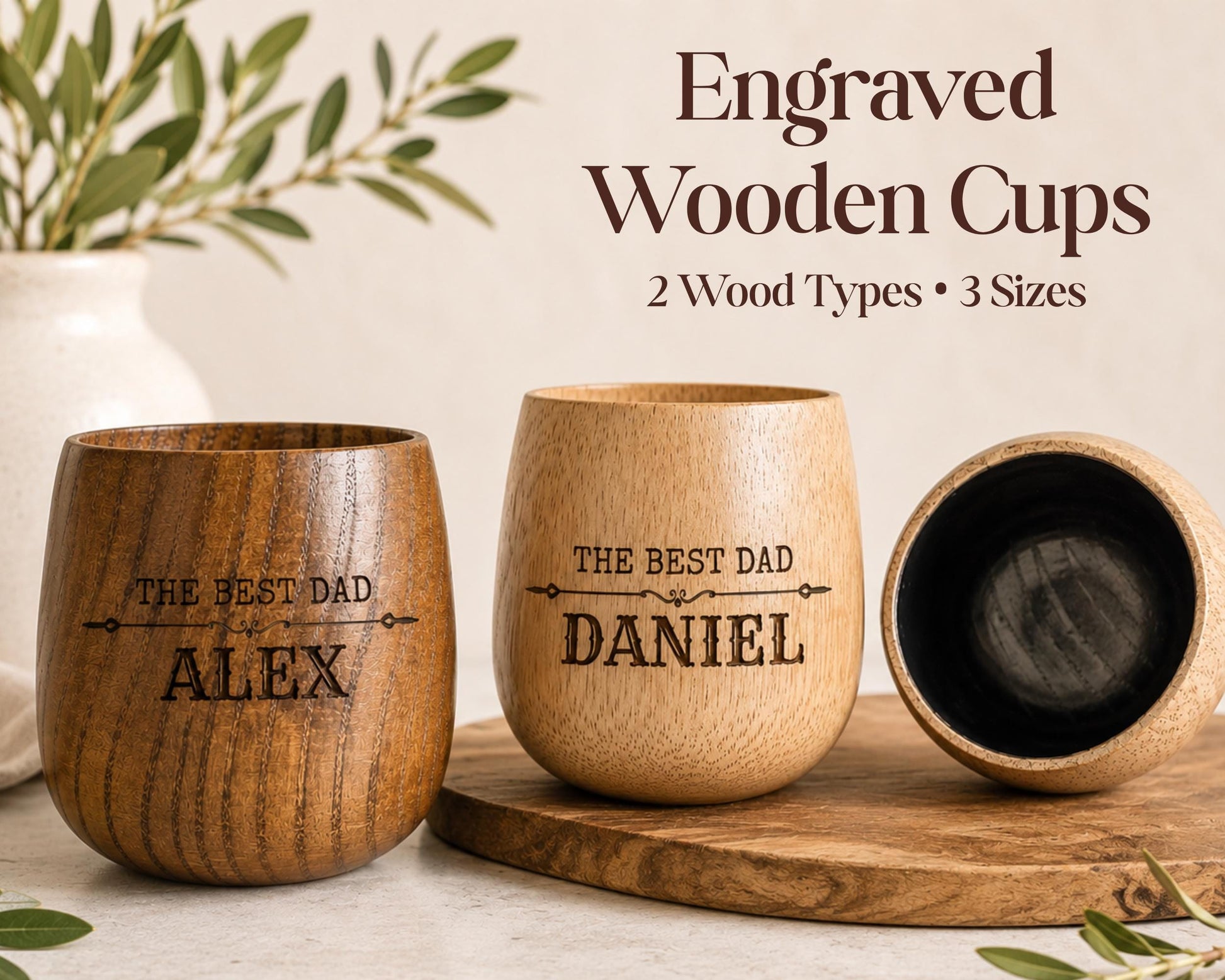 three engraved wooden cups, each with a personalized message engraved on them. The cups are made of wood and have a natural, rustic appearance. The cups are placed on a wooden surface, and there is a vase of greenery in the background. The image also includes text that reads "Engraved Wooden Cups 2 Wood Types * 3 Sizes" and "THE BEST DAD ALEX DANIEL".