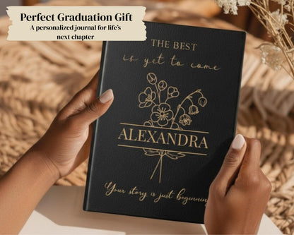 A person's hands holding a black graduation journal with gold text and a floral design.