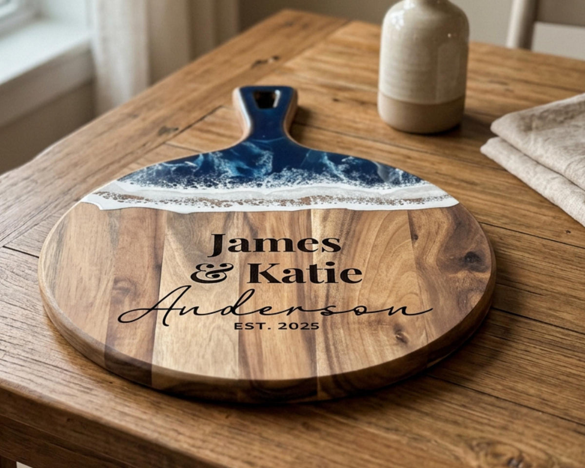A wooden cutting board with a blue and white design, featuring the names "James & Katie Anderson" and the establishment year "EST. 2025".