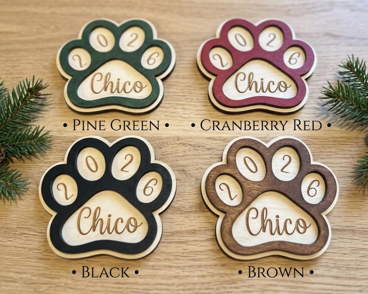 four wooden paw print ornaments, each with a different color and the year "2026" engraved on them. The paw prints are arranged on a wooden surface, with pine branches visible in the background.