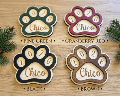 four wooden paw print ornaments, each with a different color and the year "2026" engraved on them. The paw prints are arranged on a wooden surface, with pine branches visible in the background.